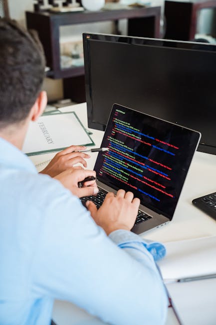 A person working on a laptop with a black screen displaying colorful lines of code, seated at a white desk in a well-lit indoor environment. The desk has a green book labeled 'Psychology' placed to th