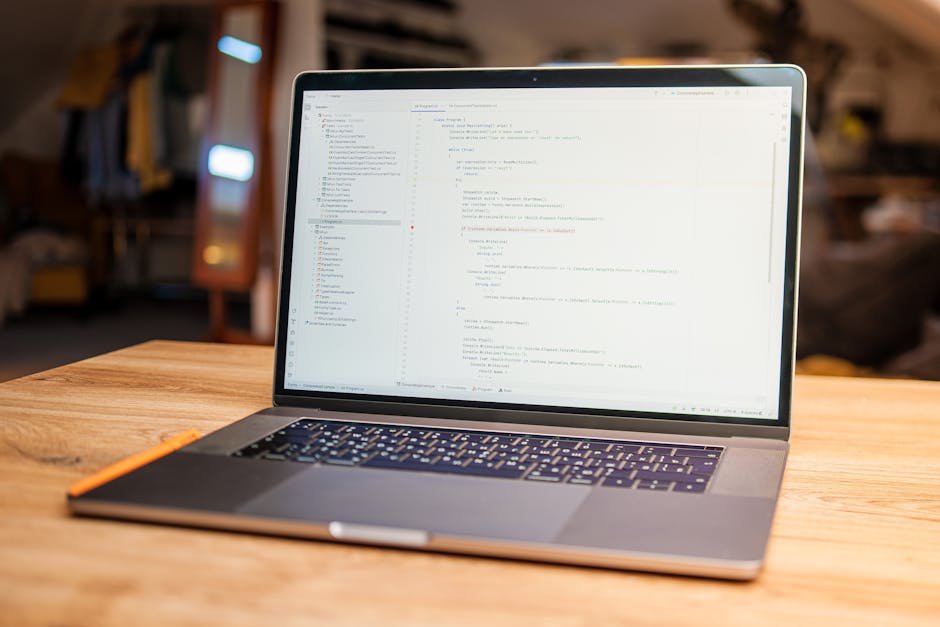 A close-up view of a silver laptop placed on a wooden table in a well-lit indoor setting, displaying lines of code on its screen. The background shows blurred elements of a cozy, rustic interior with 